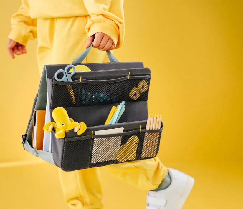 A child dressed in yellow walks across a yellow background carrying a bag containing pencils, paper, scissors and a small yellow stuffed octopus.