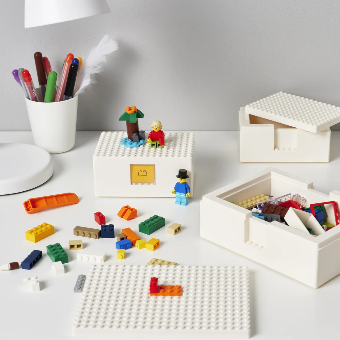 A desk with a pot of pens, some scattered lego bricks, a couple of white boxes containing more lego, and a small lego house with a lego figure in a top hat, another in a red jumper, and a small lego palm tree.